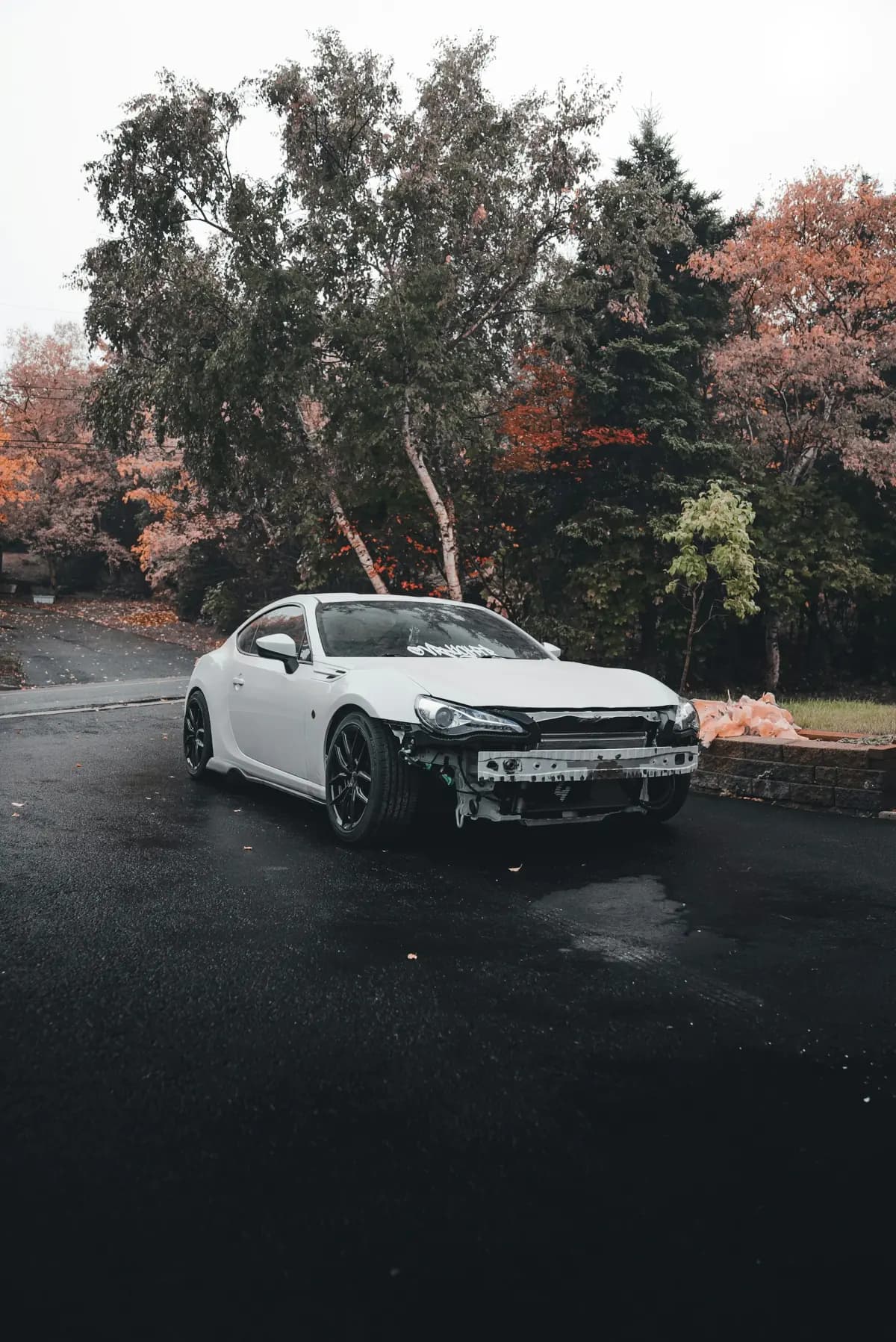 A white coupe with its front bumper torn off after a collision, parked on a wet road