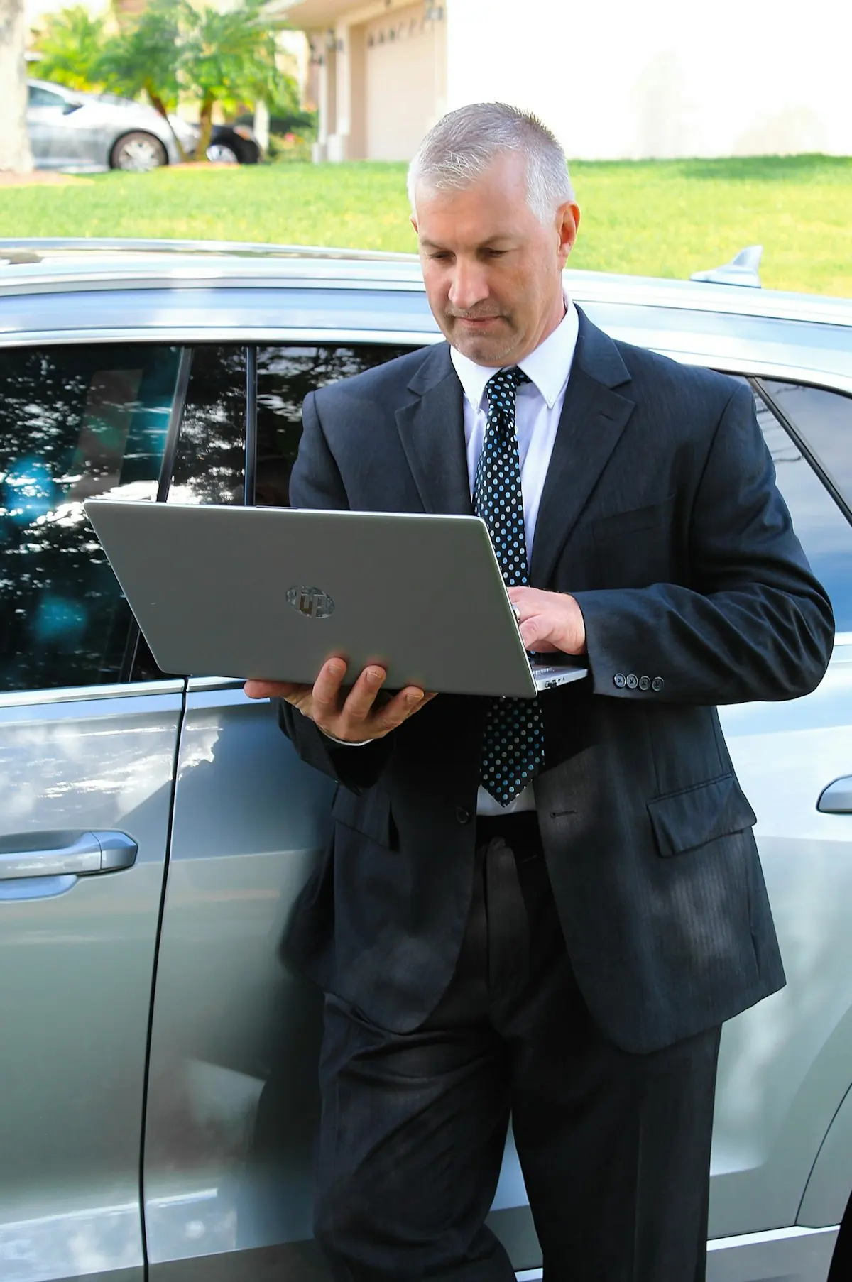 A man in a suit holding an open laptop beside a silver car