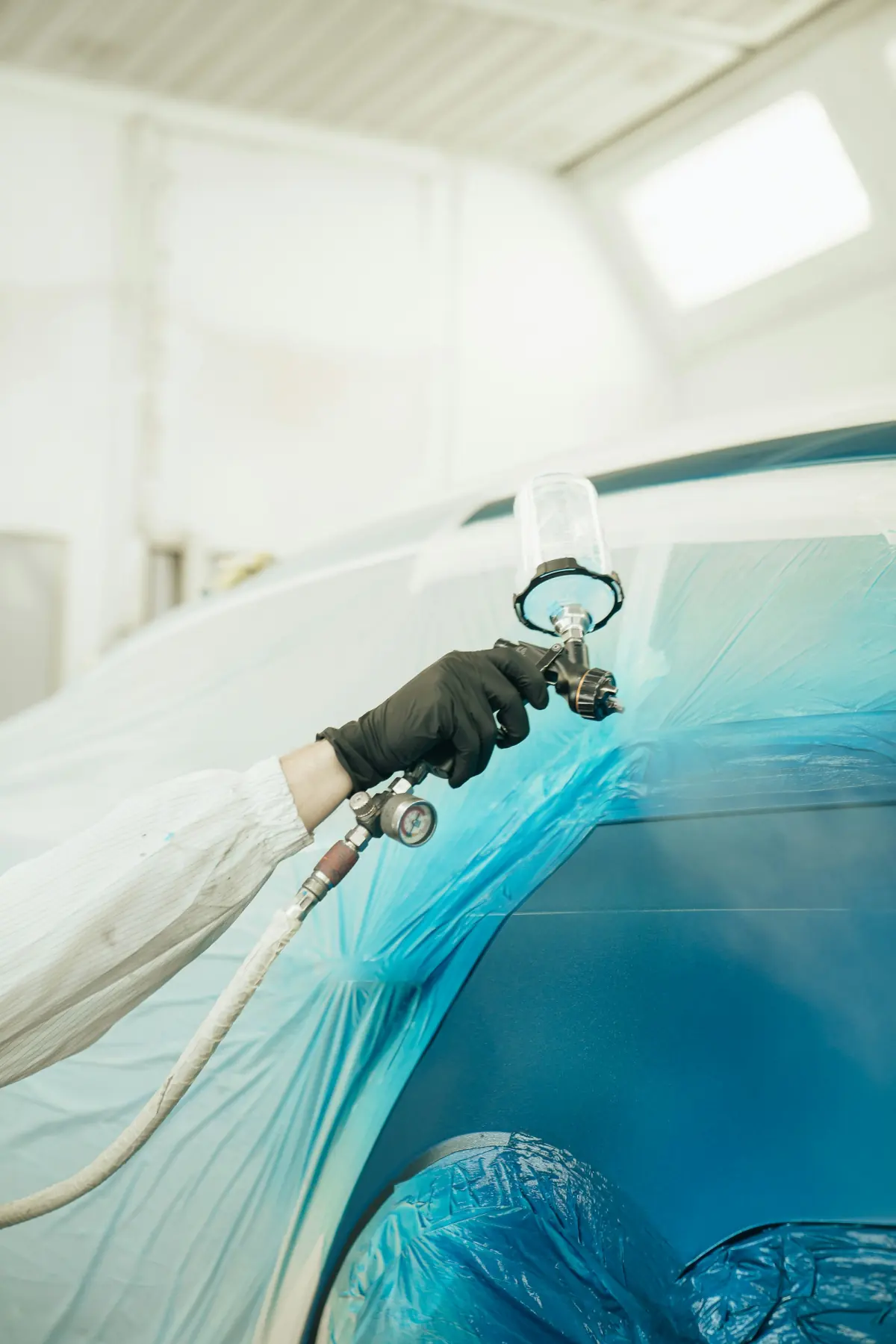 A gloved hand spraying blue paint onto a masked car body in a bright paint booth